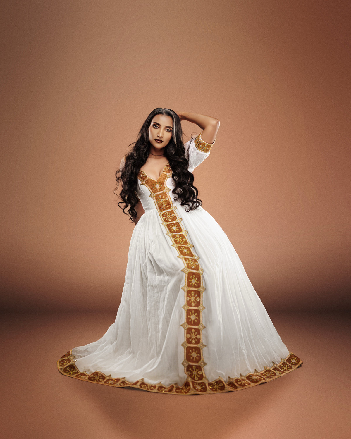 A woman modeling a long white Ethiopian national dresses with ornate gold embroidery and a gold border, posed against a solid warm brown studio background.