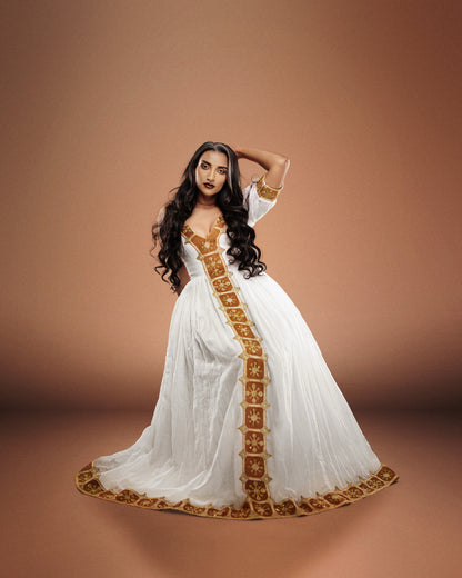 A woman modeling a long white Ethiopian national dresses with ornate gold embroidery and a gold border, posed against a solid warm brown studio background.