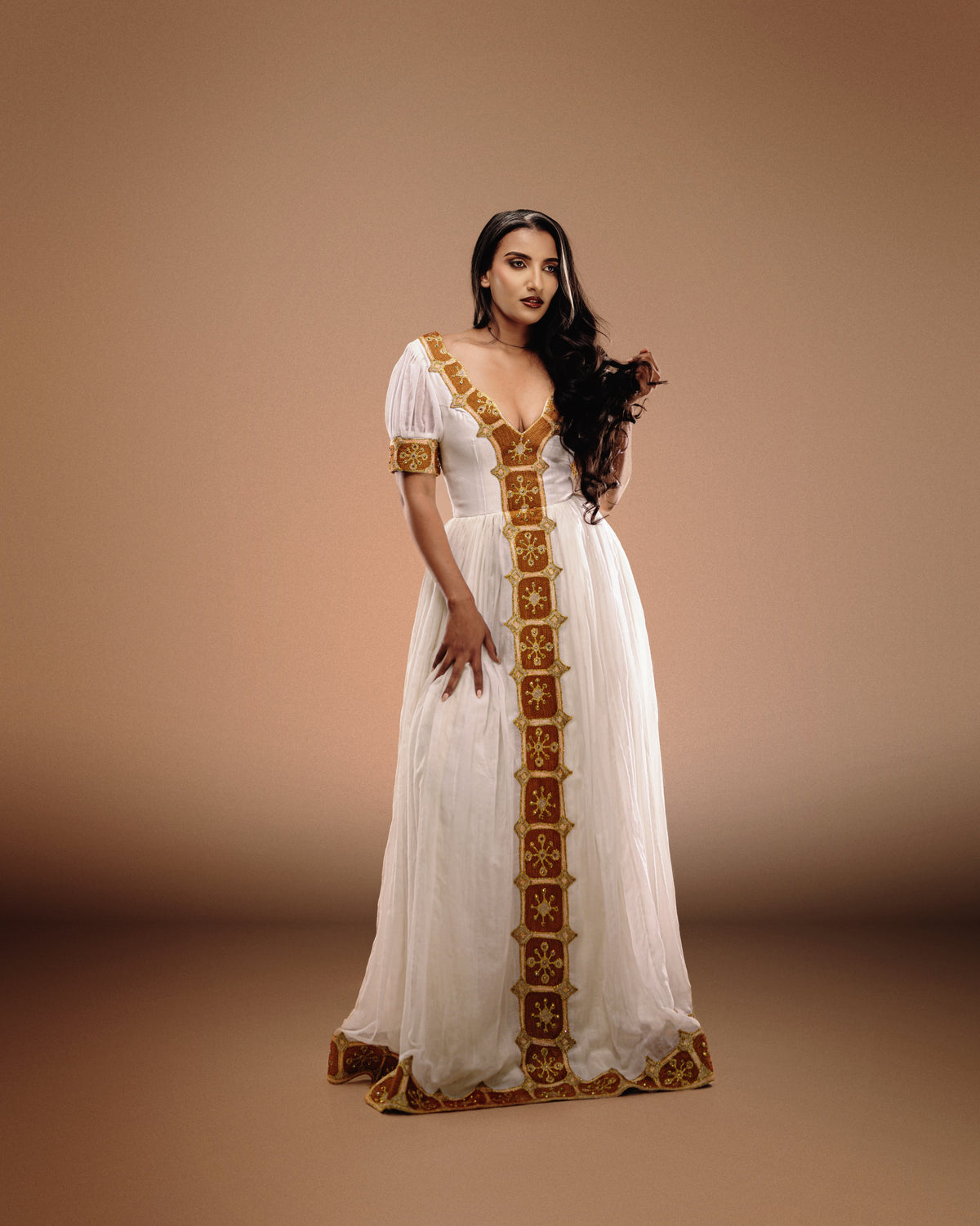 A woman modeling a long white Ethiopian national dresses with ornate gold embroidery and trim, standing against a warm beige studio backdrop.