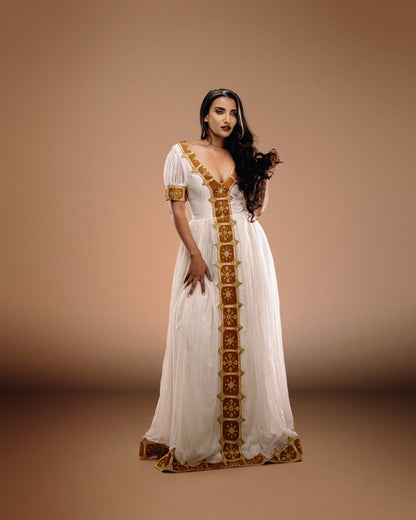 A woman modeling a long white Ethiopian national dresses with ornate gold embroidery and trim, standing against a warm beige studio backdrop.