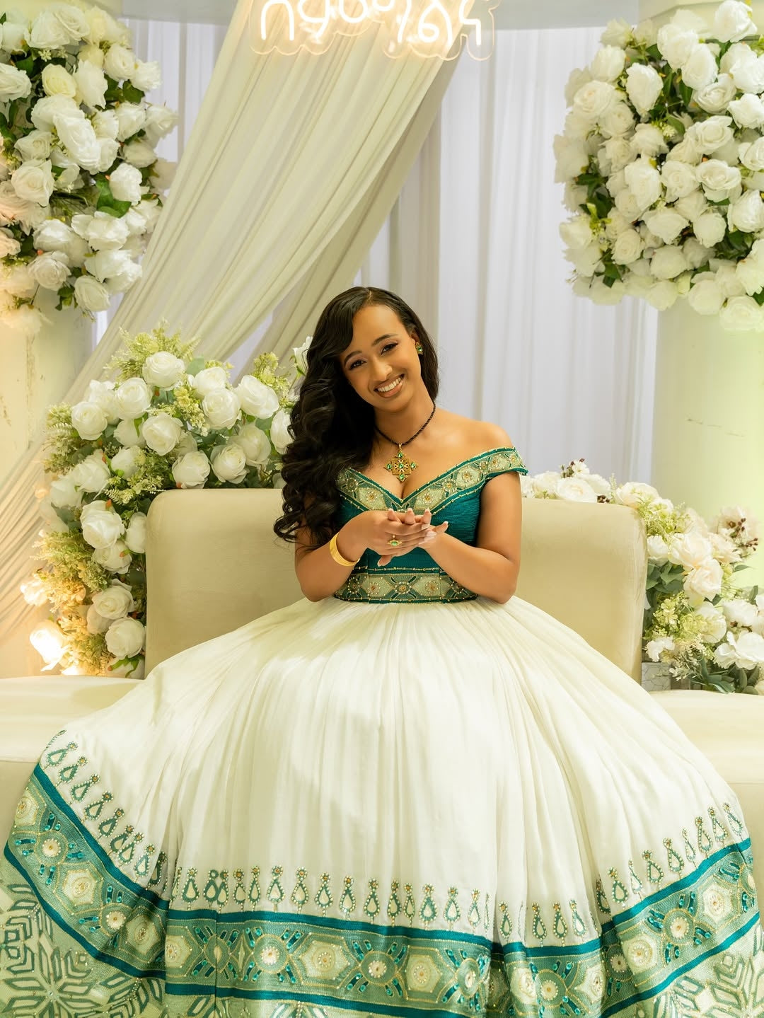 Woman in a white and green Green Habesha Wedding Kemis – sitting on a couch with floral decorations around.