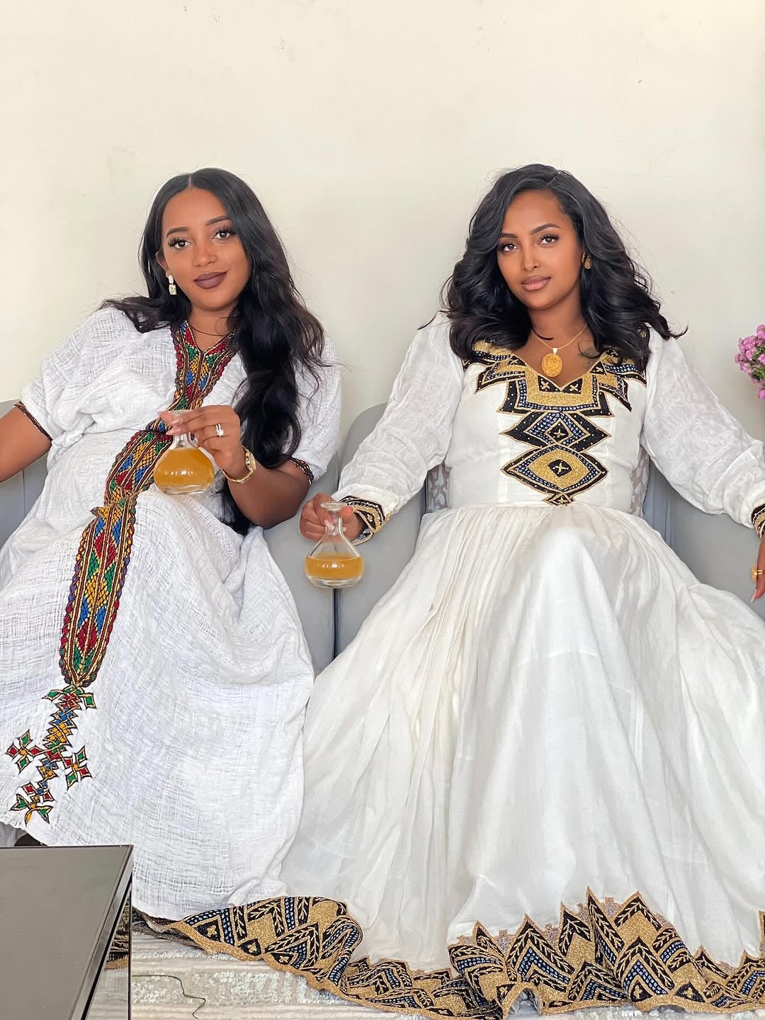 Two women in white traditional dresses with intricate patterns sitting on a couch.