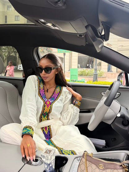 A woman wearing a white traditional long-sleeve Habesha Dress  with vibrant multicolor geometric embroidery sits inside a car. She is wearing black sunglasses and a necklace, with a Gucci handbag visible on the passenger seat next to her.