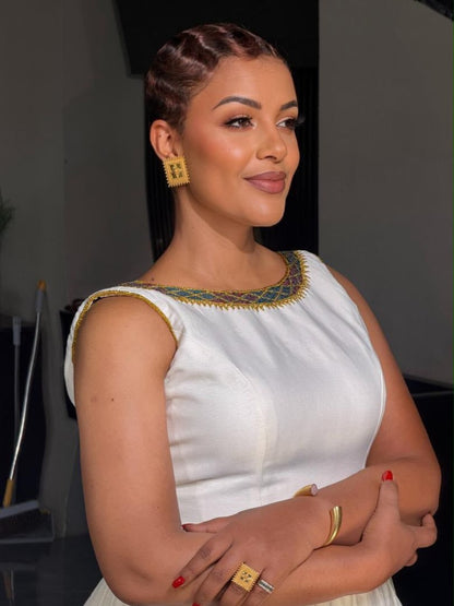 A woman wearing a sleeveless white Ethiopian National Dresses with a gold and green beaded neckline, accessorized with gold earrings, rings, and bracelets, posing with her arms crossed.