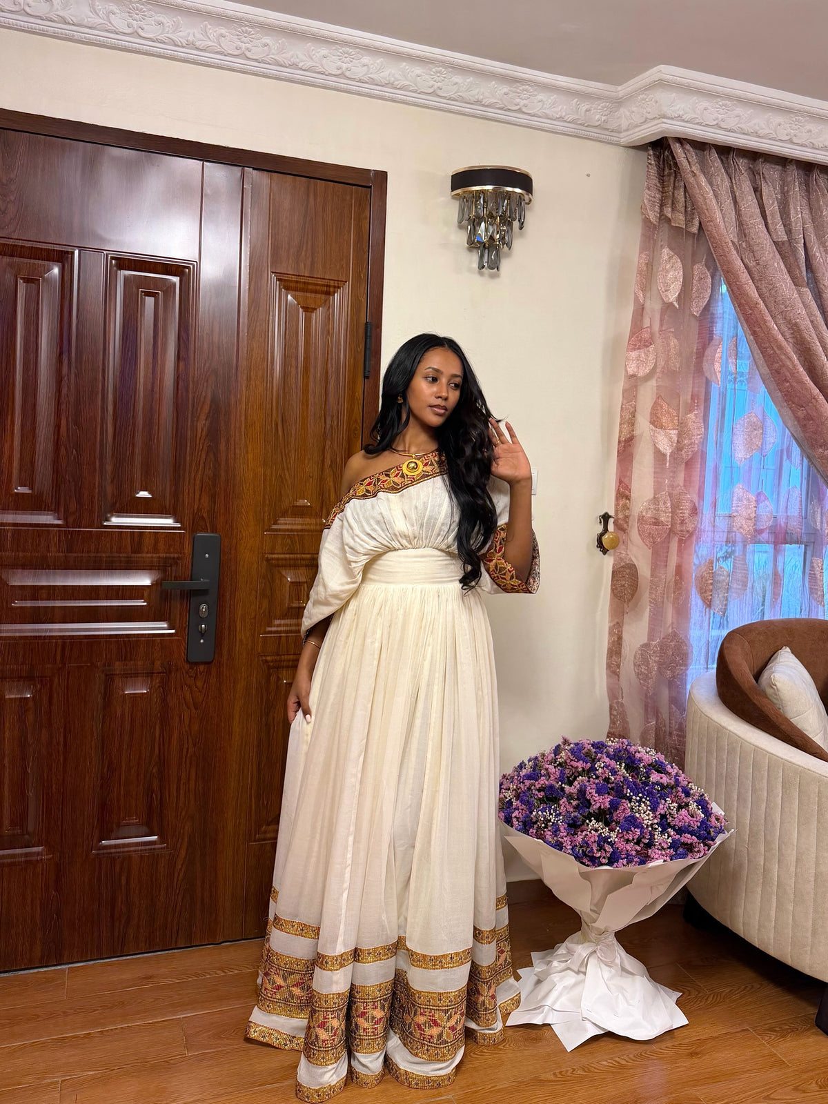 A woman wearing a long cream off-the-shoulderHabesha store  with ornate gold and brown patterns on the neckline, sleeves, and hem, standing in a decorated room next to a large bouquet of purple flowers.