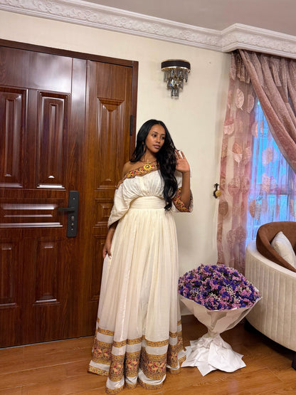 A woman wearing a long cream off-the-shoulderHabesha store  with ornate gold and brown patterns on the neckline, sleeves, and hem, standing in a decorated room next to a large bouquet of purple flowers.