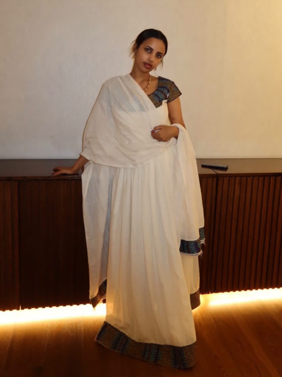 A woman wearing a white Ethiopian Clothing  with a patterned blue and black border, standing in front of a wooden counter and a plain wall.