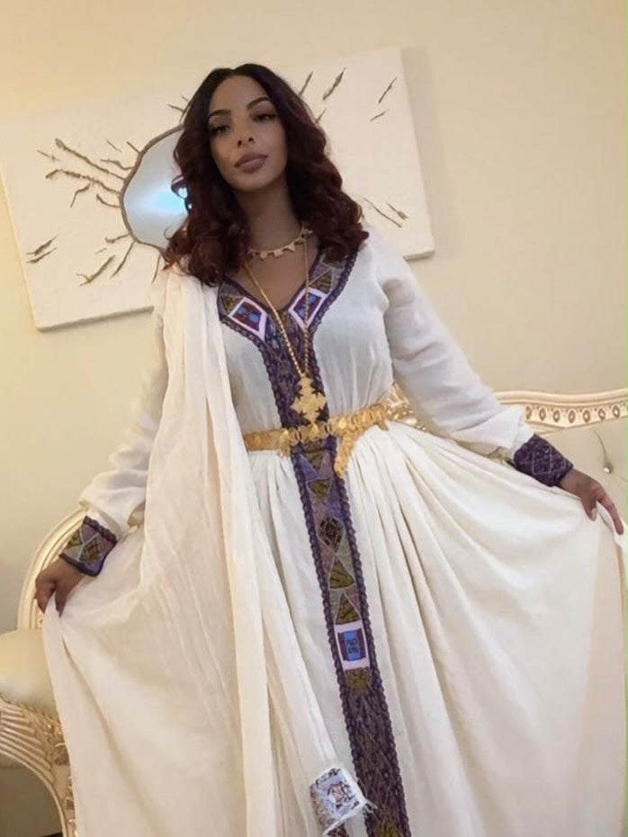 A woman wearing a traditional long white Ethiopian Shop Near Me  with purple and gold embroidered details, standing indoors and holding out the sides of the flowing garment to show its shape.