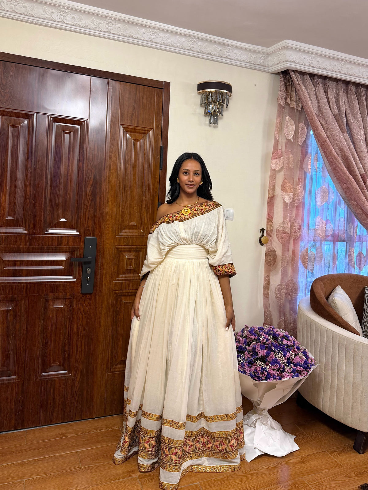 A woman stands indoors wearing a long off-the-shoulder cream Habesha store  with ornate gold and brown embroidered trim at the neckline, sleeves, and hem. The room has a dark wood door, patterned curtains, a wall sconce, and a bouquet of purple flowers near a sofa.