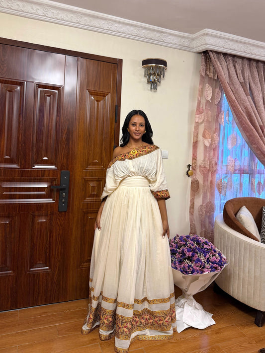 A woman stands indoors wearing a long off-the-shoulder cream Habesha store  with ornate gold and brown embroidered trim at the neckline, sleeves, and hem. The room has a dark wood door, patterned curtains, a wall sconce, and a bouquet of purple flowers near a sofa.