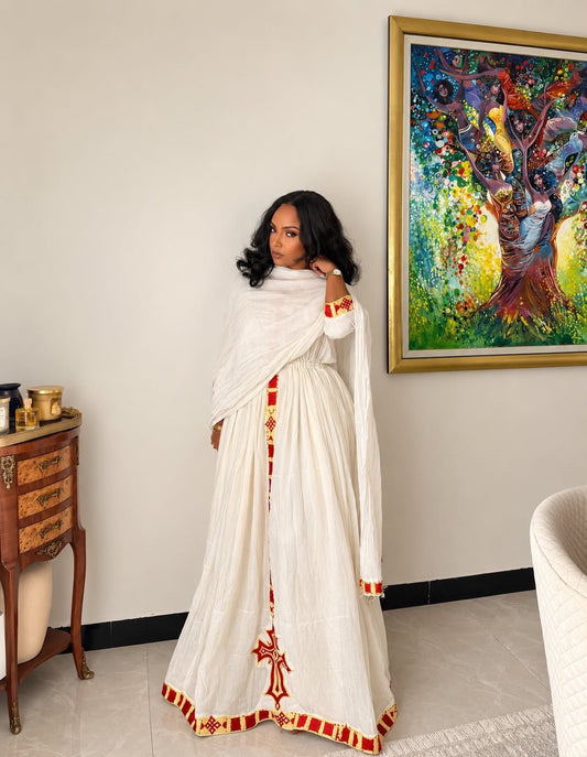 A woman wearing a traditional white embroidered Ethiopian National Dress with red and gold details, standing in a room with a framed painting on the wall.