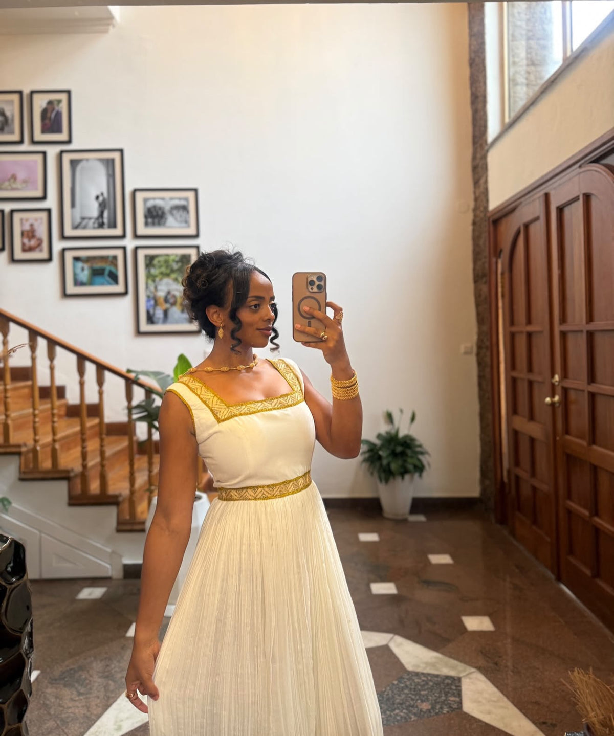 A woman takes a mirror selfie in an indoor hallway wearing a sleeveless cream-colored Habesha store with gold trim and a pleated skirt. The background features a staircase, framed pictures on the wall, and a wooden door.