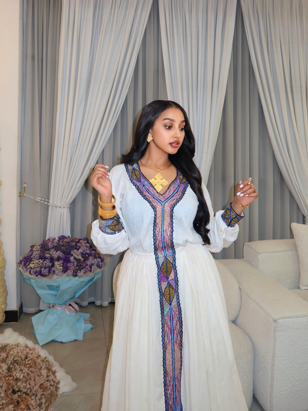 A woman wearing a white long-sleeve maxi Habesha store with colorful embroidered details on the front and cuffs, standing in front of gray curtains in an indoor setting.