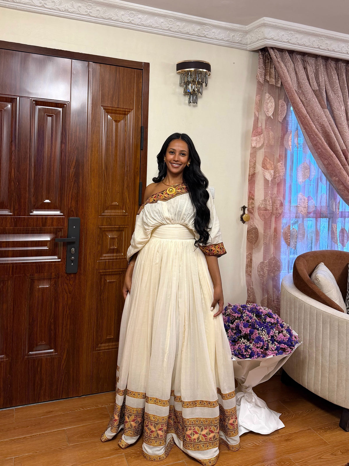 A woman wearing a cream-colored traditional Habesha store  with ornate gold and brown embroidery, standing in a decorated room with a wooden door, patterned curtains, and a floral arrangement.