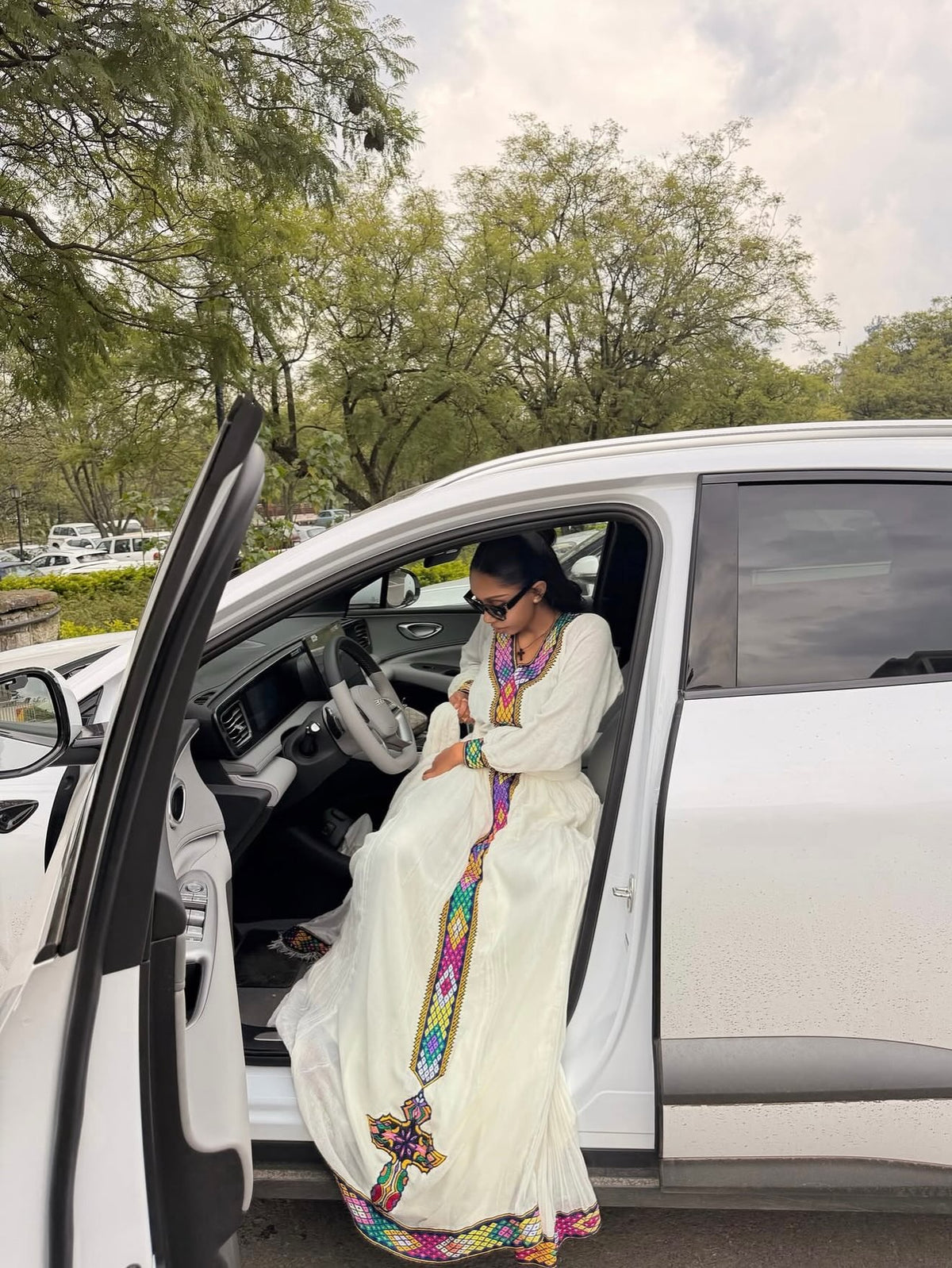 A woman wearing a long white embroidered Habesha Dress  with colorful geometric patterns sits in the open driver's side door of a white SUV outdoors.