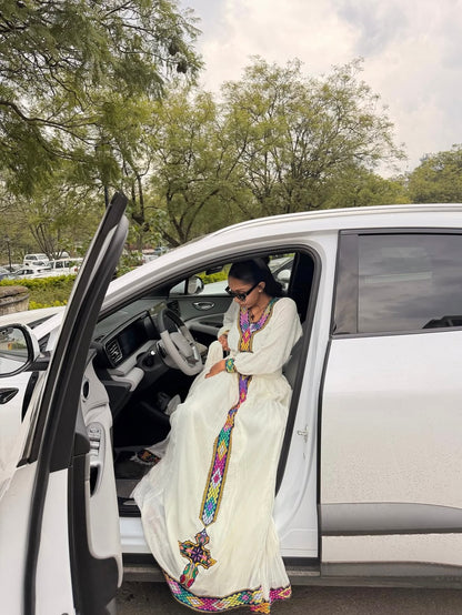 A woman wearing a long white embroidered Habesha Dress  with colorful geometric patterns sits in the open driver's side door of a white SUV outdoors.