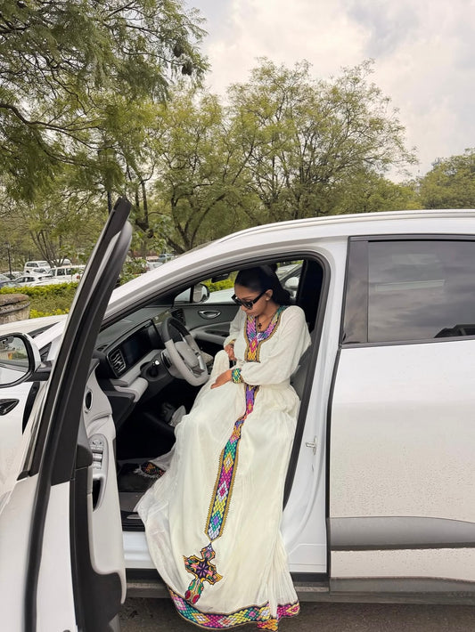 A woman wearing a long white embroidered Habesha Dress  with colorful geometric patterns sits in the open driver's side door of a white SUV outdoors.