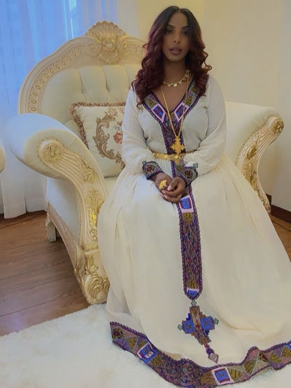 A woman wearing a traditional long white Ethiopian Shop Near Me  with purple, blue, and gold embroidered details sits on an ornate cream and gold armchair in a brightly lit room.