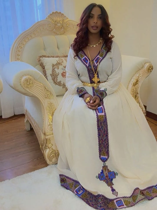 A woman wearing a traditional long white Ethiopian Shop Near Me  with purple, blue, and gold embroidered details sits on an ornate cream and gold armchair in a brightly lit room.