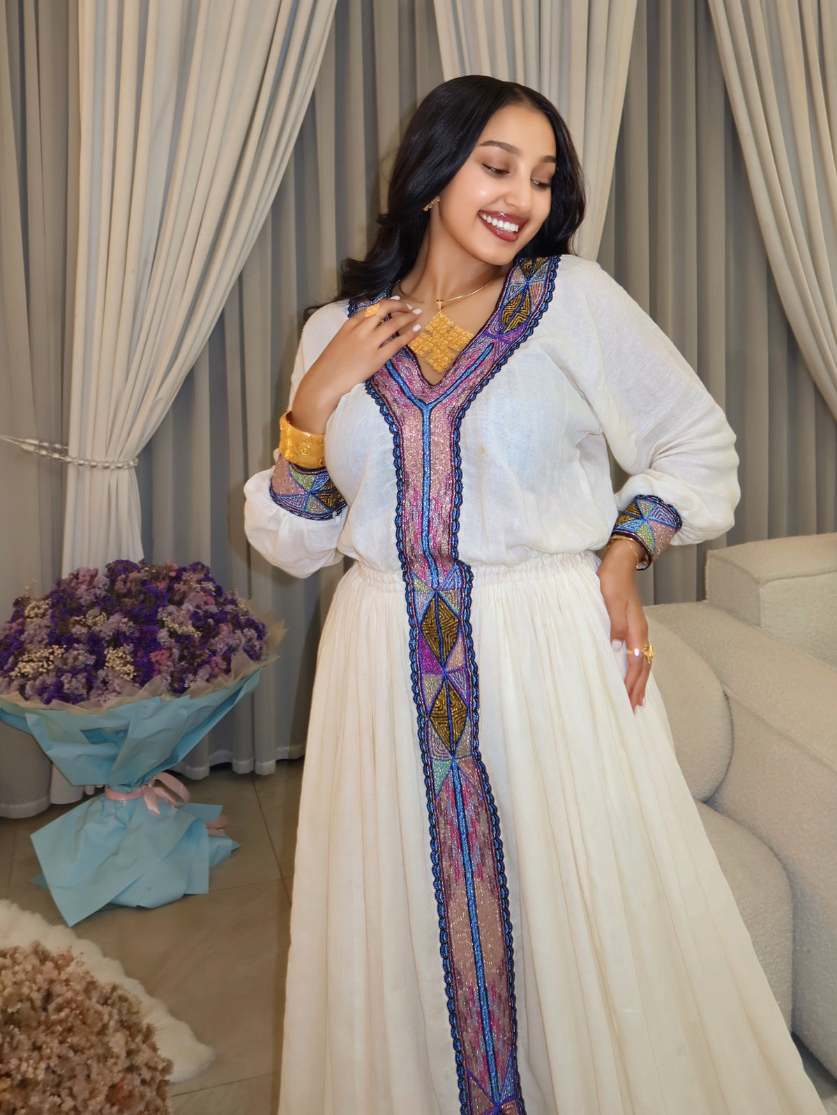 A woman wearing a white traditional long Habesha store with colorful embroidered details, standing in an indoor setting with beige curtains, a sofa, and a bouquet of purple flowers.