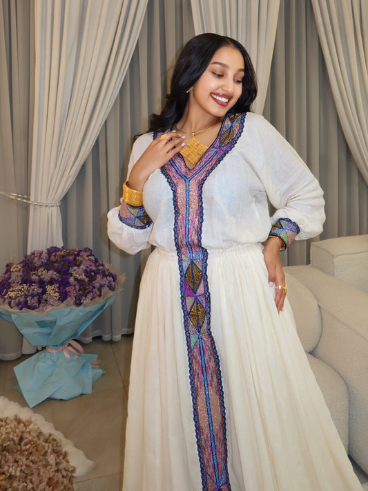 A woman wearing a white traditional long Habesha store with colorful embroidered details, standing in an indoor setting with beige curtains, a sofa, and a bouquet of purple flowers.