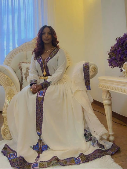 A woman wearing a traditional white embroidered Ethiopian Shop Near Me  with purple and gold detailing, seated on an ornate white armchair in an indoor setting.