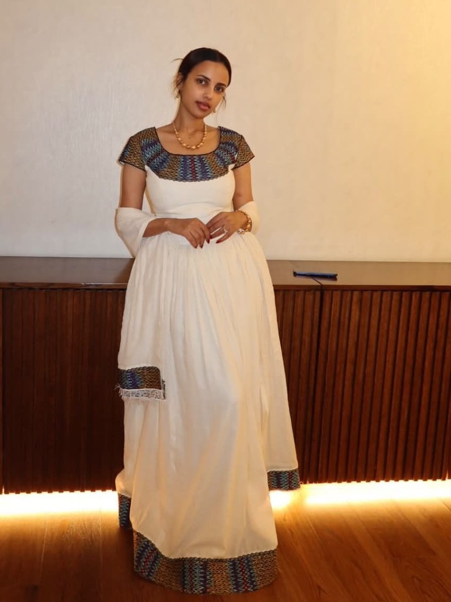 A woman wearing a white traditional Ethiopian Clothing  with multicolor patterned embroidery on the neckline, sleeve cuffs, and hem, standing in front of a wooden slatted wall with warm lighting.
