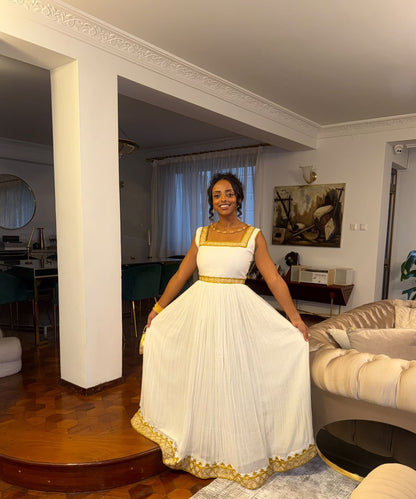 A woman wearing a white sleeveless maxi Habesha store with gold trim and a gold sash, holding out the skirt to show its full volume, standing in a living room.