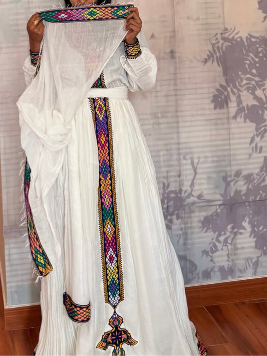 A person wearing a long white traditional Habesha Dress  with colorful embroidered borders and holding a matching sheer white dupatta in front of their face. The dress features intricate geometric multicolor embroidery along the waist, hem, and cuffs.
