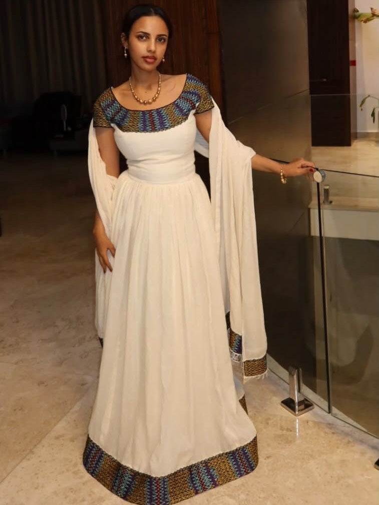A woman wearing a white floor-length Anarkali Ethiopian Clothing  with a heavily embroidered multicolor neckline and a matching patterned border along the hem, standing indoors near a glass railing.
