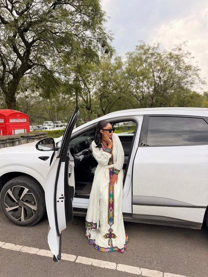 A woman wearing a long white embroidered Habesha Dress  and sunglasses stands beside an open white car door outdoors, with trees and a red kiosk in the background.