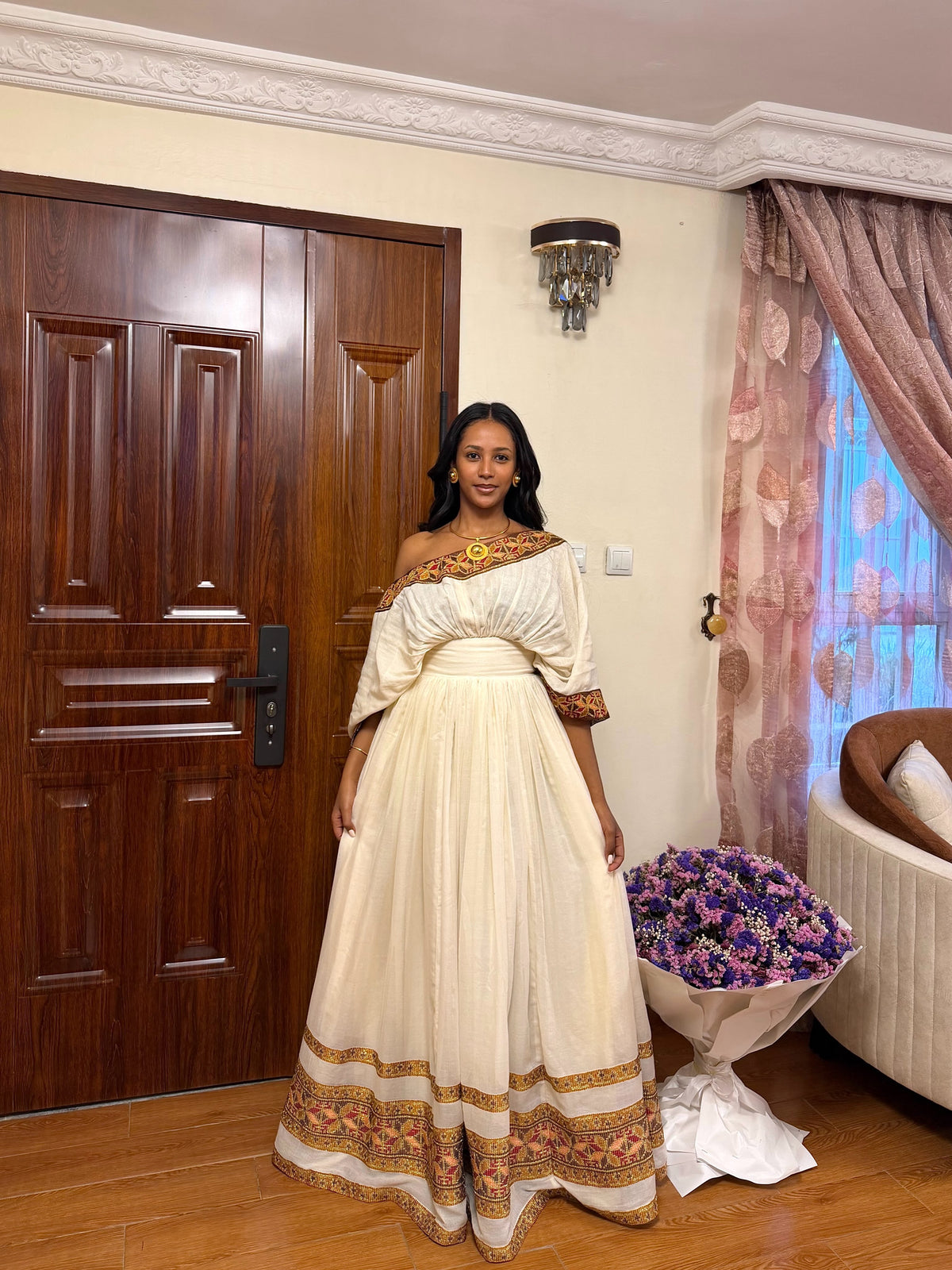A woman stands indoors wearing a voluminous off-the-shoulder cream Habesha store  with ornate gold and brown embroidery concentrated on the bodice, sleeves, waistband, and hem. The room has a dark wood door, patterned curtains, a wall sconce, and a bouquet of purple flowers on the floor.