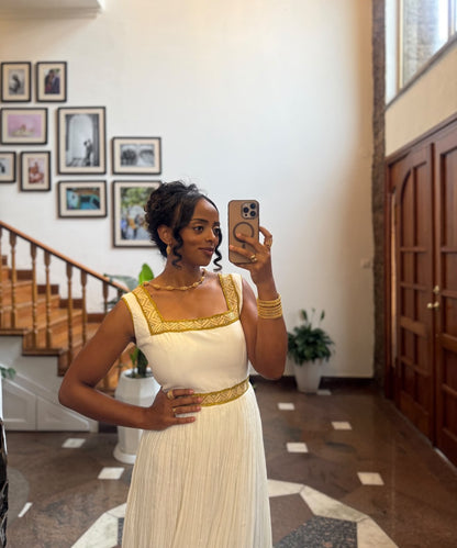 A woman takes a mirror selfie in a grand indoor hallway, wearing a white sleeveless Habesha store with gold trim at the neckline and waist, and holding a gold-rimmed smartphone.