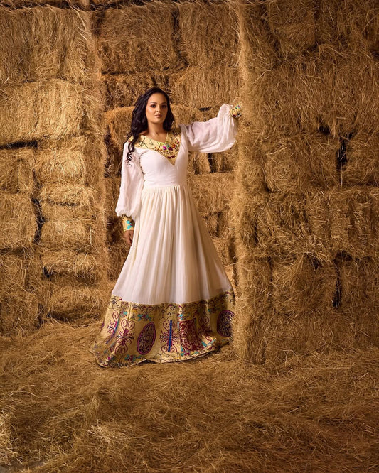 Woman in a white Ethiopian cultural dress with colorful patterns standing between two hay bales.