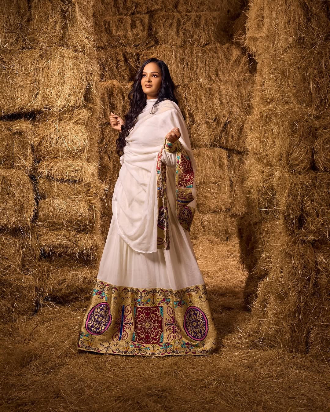 Woman in a white and patterned Ethiopian traditional dress outfit standing in a hay barn.