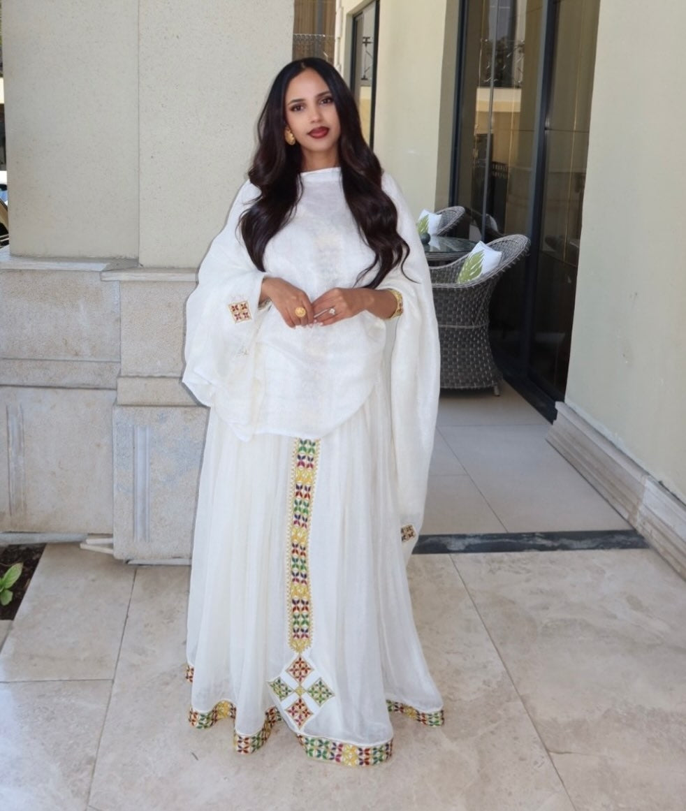 Woman in a white traditional Ethiopian cultural dress with colorful patterns standing in an outdoor setting.