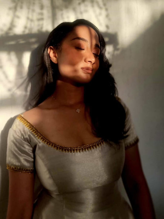 Woman in a light gray Ethiopian cultural dress with gold accents against a textured wall.