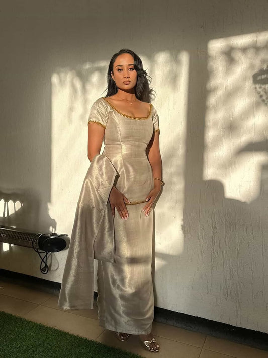 Woman in a silver Ethiopian cultural dress  standing against a white wall with a bench.