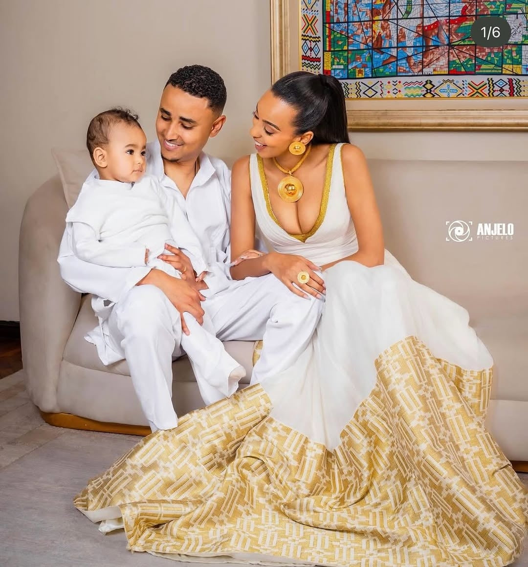 Family of three sitting together on a couch, with a woman in a white and gold dress and a man holding a child.