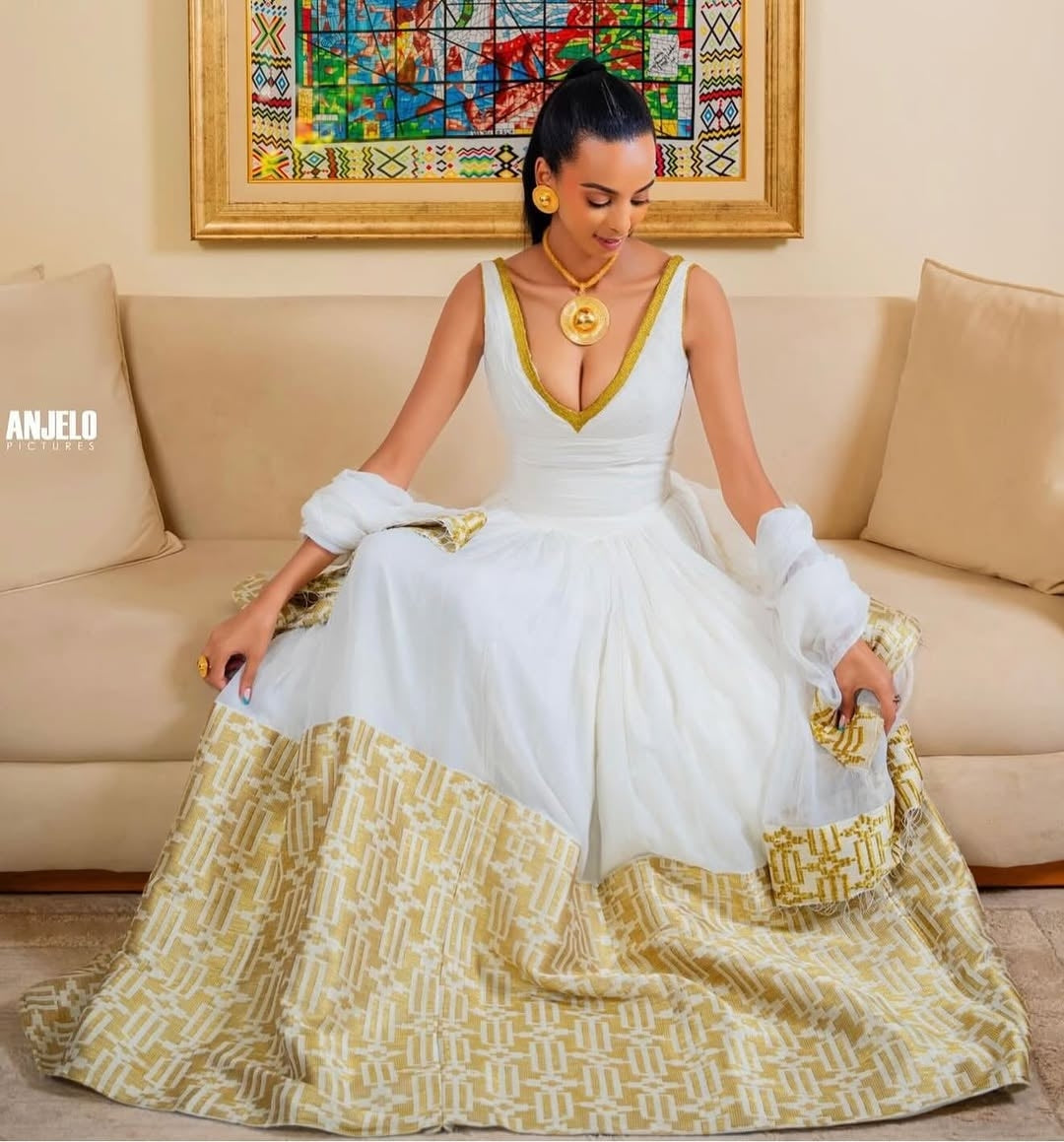 Woman in a white and gold habesha dress sitting on a decorative chair in an elegant interior setting.