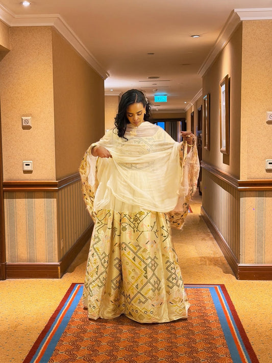 Woman holding a white  Ethiopian traditional dress over a embroidered outfit in a hallway.