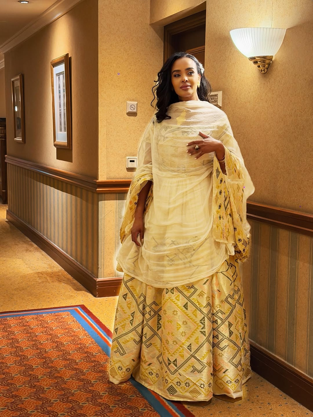 Woman in a yellow  Ethiopian traditional dress outfit standing in a hallway with beige walls and a patterned rug.