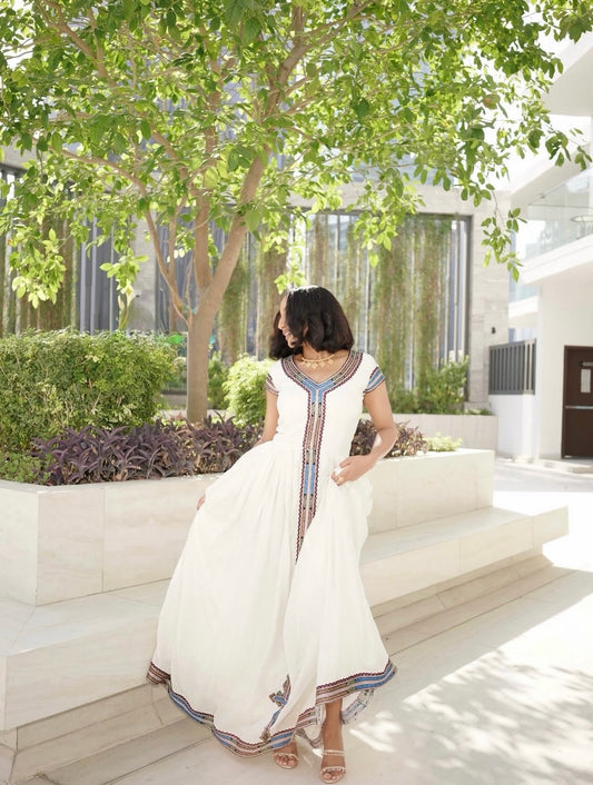 A woman wearing a white maxi Ethiopia clothes with colorful embroidered details stands on outdoor stone steps under a leafy tree.