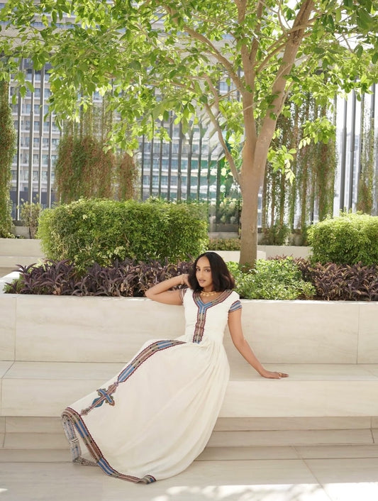 A woman wearing a long white Ethiopia clothes with colorful embroidered details sits on a light-colored outdoor stone bench in a garden setting with trees and plants.