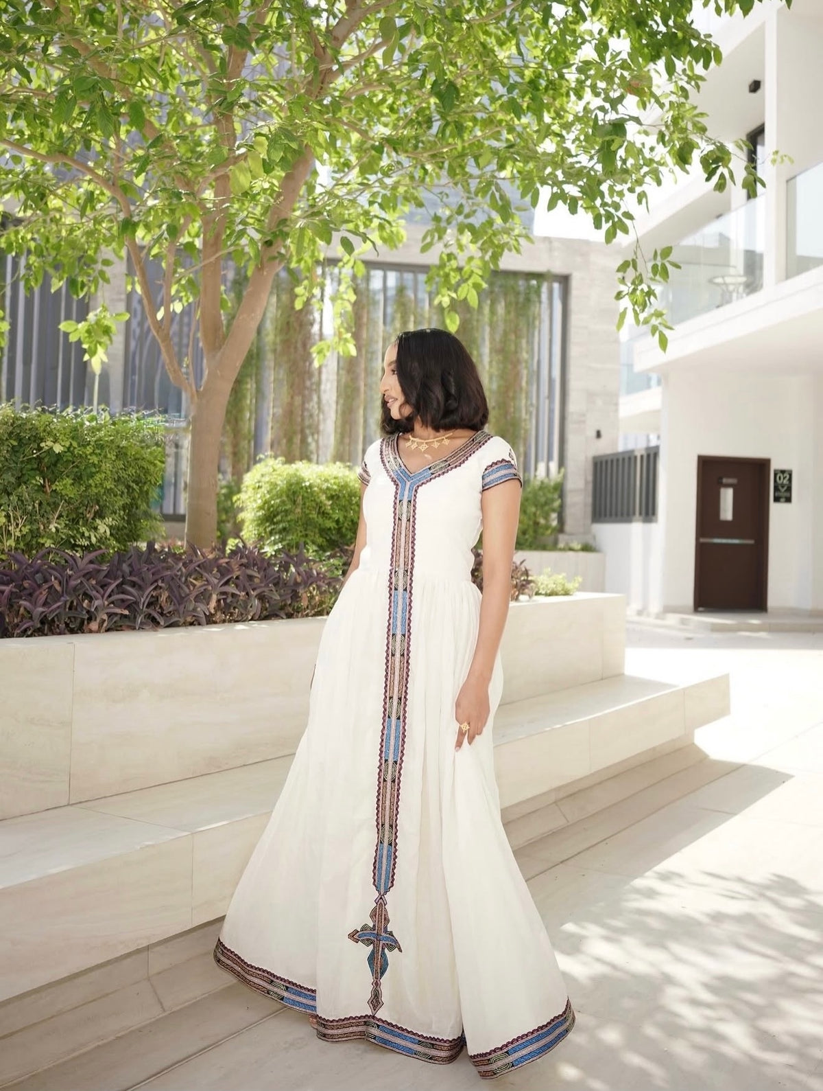 A woman wearing a long white Ethiopia clothes with colorful embroidered details along the front and hem, standing outdoors in front of a modern building with trees and stone steps.