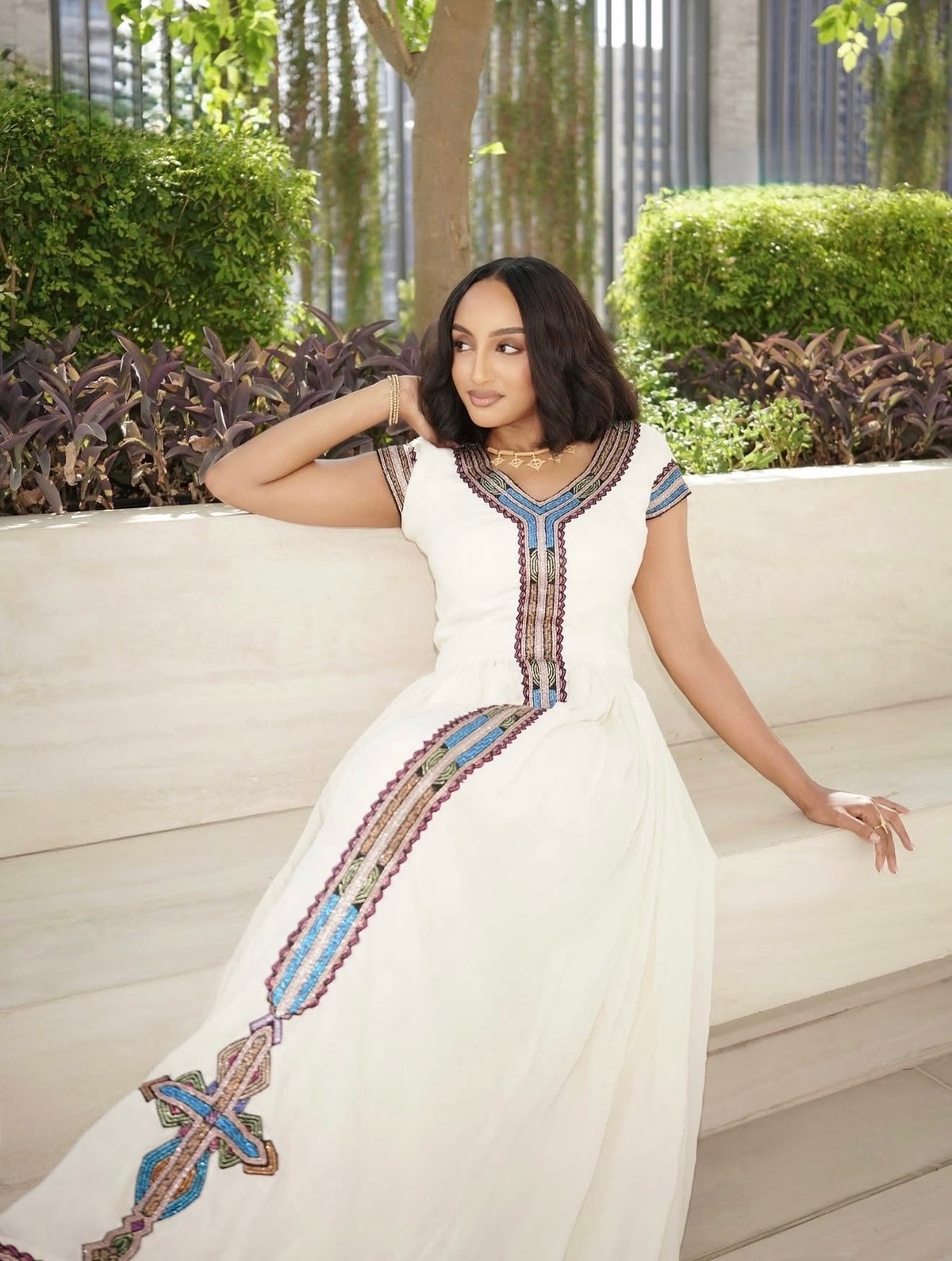 A woman wearing a white Ethiopia clothes with colorful geometric embroidery, sitting on a light-colored stone bench in an outdoor garden setting.
