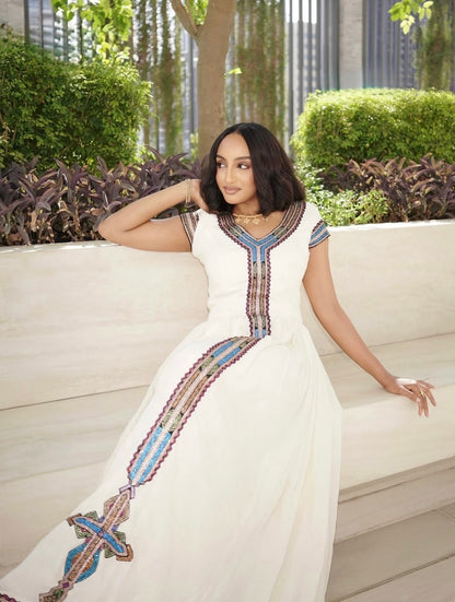 A woman wearing a white Ethiopia clothes with colorful geometric embroidery, sitting on a light-colored stone bench in an outdoor garden setting.