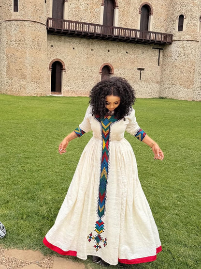 Woman in a long  Ethiopian Libs with colorful embroidery standing on grass in front of a castle.