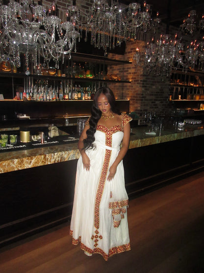 A woman wearing a white traditional Habesha Kemis with orange and gold embroidery, standing in front of a dimly lit bar with hanging crystal chandeliers.