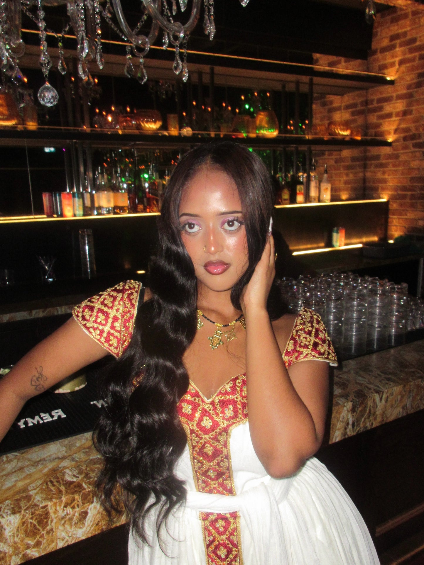 A woman posing in a white dress with red and gold embroidered Habesha Kemis, set against a dimly lit bar background.